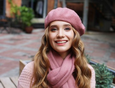 Uzun kıvırcık saçlı, pembe günlük kıyafetler, atkı ve şapka giymiş, açık hava portresi olan genç bir kadın..