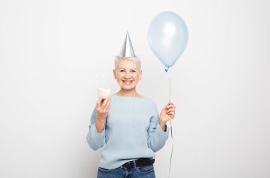 Mutlu yaşlı kadın koni şapkası takıyor doğum gününü mum ve gri arka planda mavi balonla kutluyor.