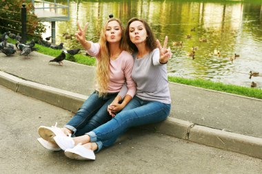 Two women hugging while sitting next to the lake. Mother and daughter, happy and beautiful. They pose and show the sign of victory. Summer day in the park.
