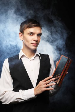 Stüdyoda poz veren profesyonel makyöz erkek, palet ve fırça tutuyor. Dumanlı karanlık bir arka plan. Moda konsepti.