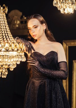 Şık bir avizesi olan güzel bir odada, gece elbisesi ve eldivenleri içinde büyüleyici bir kadın. Moda konsepti.
