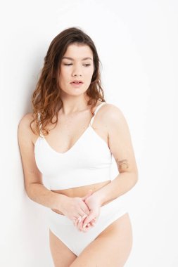 Uzun siyah kıvırcık saçlı, beyaz iç çamaşırı giyen, beyaz arka planda poz veren bir manken.