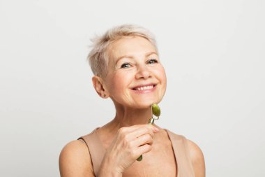 60 'lı yaşlarda güzel bir kadın. Yüz masajı yapıyor. Gri arka planda yeşil kuvars taşı kullanıyor..