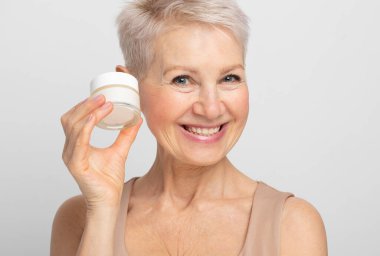 Kısa saç stili gülümseyen ve elinde bir kavanoz krema tutan çekici, yaşlı bir kadın..