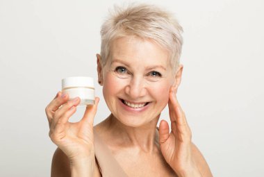 A charming elderly woman with a short hairstyle smiles and holds a jar of cream in her hands. clipart