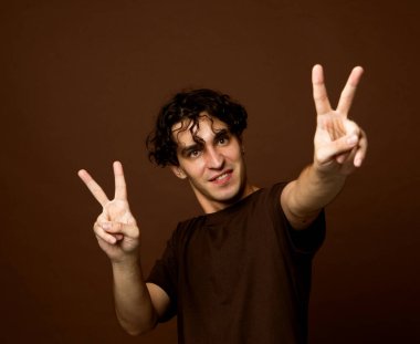 Genç kıvırcık adam kahverengi arka planda zafer işareti yapıyor..