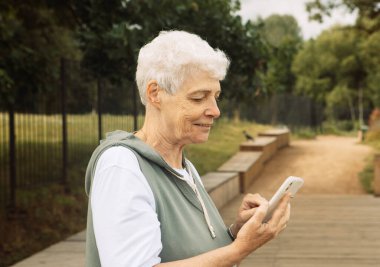 Yaşlı mutlu kadın emekli elinde akıllı bir telefonla yaz parkında mesaj yazıyor.