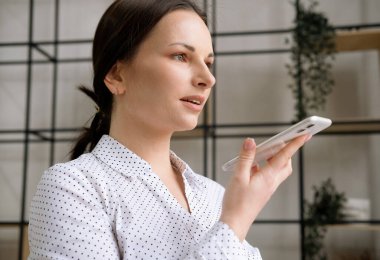 Beyaz bluzlu genç bir kadın ofisteki bir akıllı telefondan sesli mesaj kaydediyor. İş ve insan kavramı.