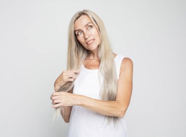 Güzellik ve yaşlılar konsepti: Güzel yaşlı kadın açık gri arka planda uzun gri saçlara dokunur. Kapat..