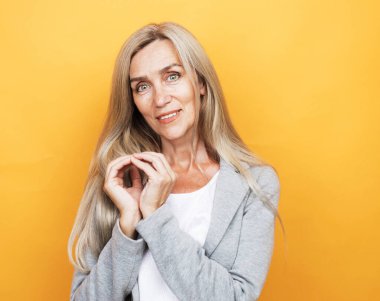 Mükemmel, hoş, yaşlı, yaşlı, uzun saçlı, sarı arka planda parmakları olan kalp figürünü gösteren güzel bir kadın..