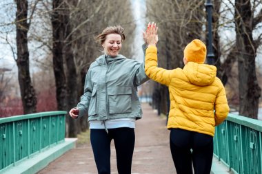 İki gülümseyen bayan arkadaş parkta koşuyor, birbirlerine beşlik çakmaya doğru koşuyorlar..