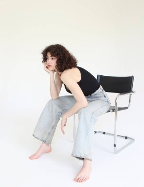 Kot pantolonlu ve beyaz tişörtlü güzel bir kadın model beyaz arka planda bir sandalyede oturuyor..