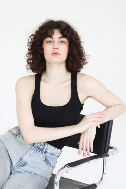 Kot pantolonlu ve beyaz tişörtlü güzel bir kadın model beyaz arka planda bir sandalyede oturuyor..