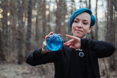 Mavi saçlı genç bir kadın ormanda mavi bir kristal küre tutuyor. Sahne gizemli ve sakin.