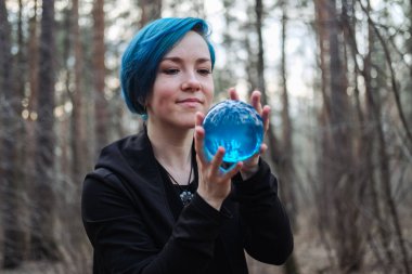 Mavi saçlı bir kadın ormanda mavi bir kristal küre tutuyor. Sahne gizemli ve sakin.