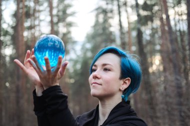 Mavi saçlı genç bir kadın ormanda mavi bir kristal küre tutuyor. Sahne gizemli ve sakin.