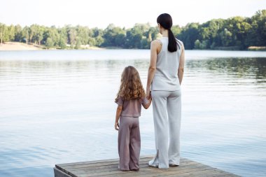 Genç bir anne ve altı yaşındaki kızı gölün yakınındaki bir rıhtımda durup denize bakıyorlar, arkadan görüyorlar..