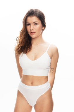 Uzun kıvırcık saçlı, beyaz iç çamaşırlı, beyaz arka planda stüdyoda poz veren genç bir kadın model.