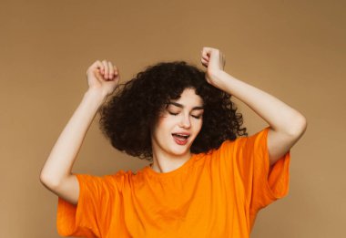 Kıvırcık saçlı, gülümseyen genç kadın bej renkli arka planda yalnız dans ediyor..