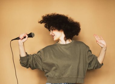 Neşeli, kıvırcık saçlı genç bir kadın mikrofona şarkı söylüyor ve dans ediyor..