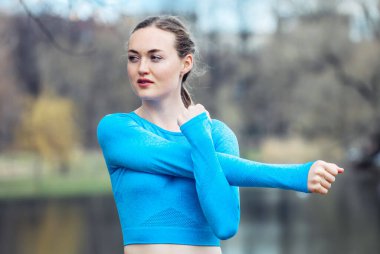 Mavi spor kıyafetli genç bir kadın bahar günü göl kenarında kol egzersizi yapıyor..