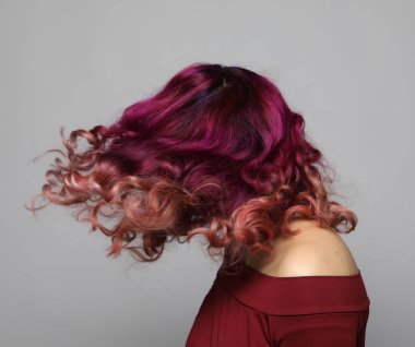 Çok renkli kıvırcık saçlı, saçlarını sallayan, saçlarında rüzgar olan genç bir kadın. Gri arkaplandaki portre. Kapat..