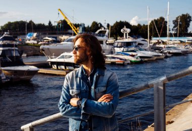 Uzun saçlı, güneş gözlüğü takan çekici bir adam tekne ve yatların yakınındaki sette yürüyor. Yaz günü. Moda ve tarz.