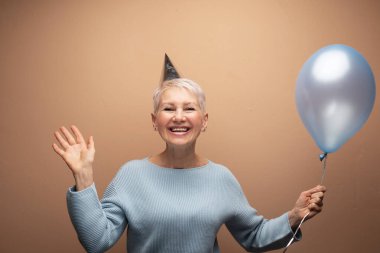 Doğum gününü kutlayan konik şapka takan güzel, yaşlı bir kadın. Bej arkaplan üzerinde portre.