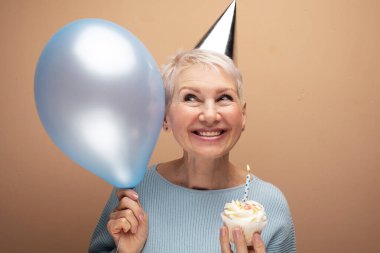 Doğum gününü kutlayan konik şapka takan güzel, yaşlı bir kadın. Bej arkaplan üzerinde portre.