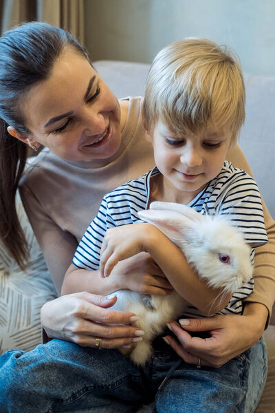 Шестилетний мальчик и его мать играют с белым кроликом дома.