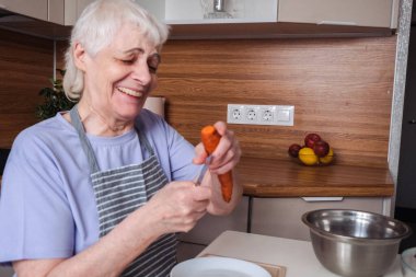 Neşeli yaşlı emekli kadın salata için havuç soyar ve güler..