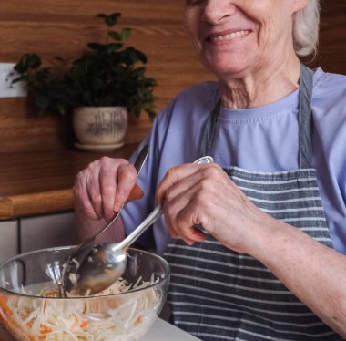 Kısa saçlı yaşlı bir kadın mutfak tezgahında taze lahana ve havuç salatası hazırlıyor. Resmi kapat.