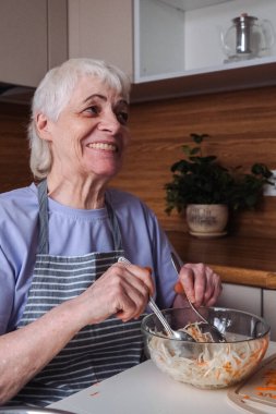 Kısa saçlı yaşlı bir kadın mutfak tezgahında taze lahana ve havuç salatası hazırlıyor. Resmi kapat.