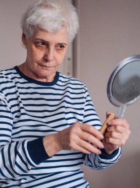 Güzel, yaşlı bir kadın ayna tutuyor ve evde yüz kremi sürüyor. Yaşam tarzı kavramı.
