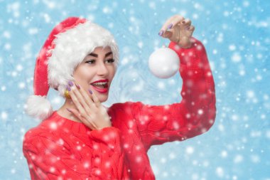 Kırmızı Noel Baba şapkalı güzel bir kadın portresi ve kar tanelerinin arkasında beyaz bir Noel topu tutan kırmızı örgü süveter. Yeni yıl konsepti.