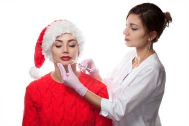 Doktor kozmetik uzmanı Noel Baba şapkalı güzel bir kadının dudaklarına iğne yapıyor. Yeni Yıl ve Kozmetoloji Konsepti