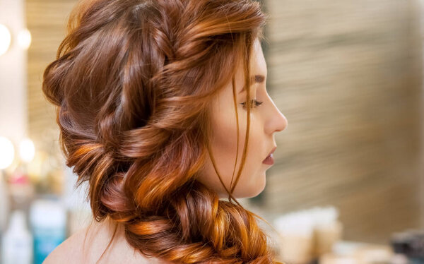 Beautiful, with long, red-haired hairy girl, hairdresser weaves a French braid, in a beauty salon. Professional hair care and creating hairstyles.