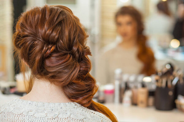 Beautiful, with long, red-haired hairy girl, hairdresser weaves a French braid, in a beauty salon. Professional hair care and creating hairstyles.