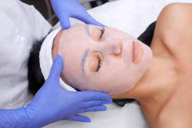 Güzellik salonundaki genç bir kadının yüzüne çarşaf maskesi takmak suretiyle cildi temizleme ve nemlendirme prosedürü. Kozmetoloji ve profesyonel cilt bakımı..