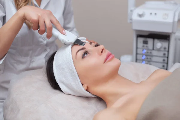 The doctor-cosmetologist makes the ultrasound cleaning procedure of the facial skin of a beautiful, young woman in a beauty salon. Cosmetology and professional skin care.