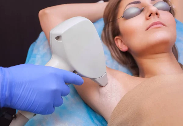 The cosmetologist does the laser hair removal procedure in the armpit zone, to a young woman in a beauty salon.