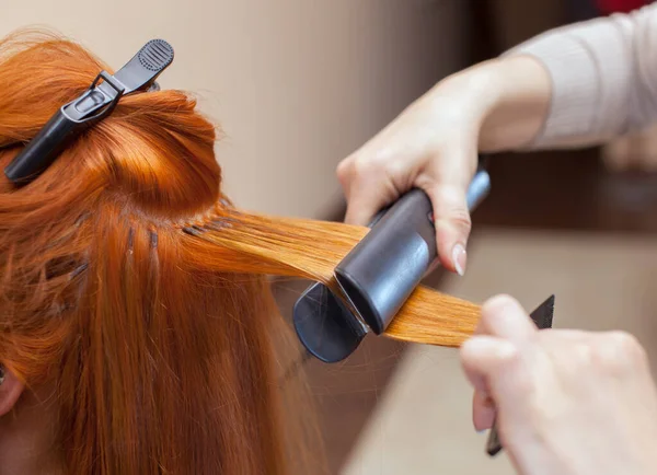 The hairdresser does hair extensions to a young girl, a redhead girl in a beauty salon. Professional hair care.