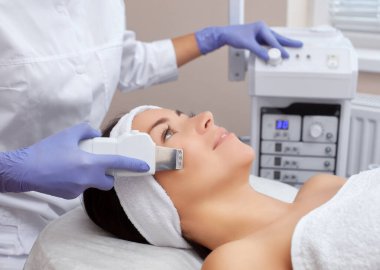 Doktor-kozmetik uzmanı güzellik salonundaki genç ve güzel bir kadının yüz derisinin ultrason temizleme prosedürünü uygular. Kozmetoloji ve profesyonel cilt bakımı.