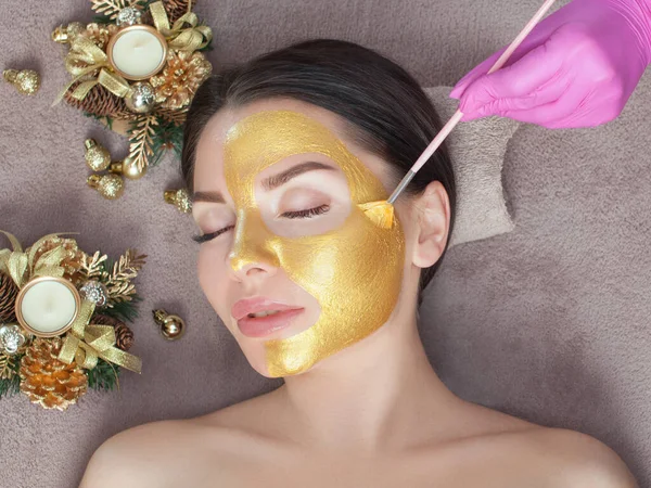 Beautician makes a golden mask to rejuvenate the skin to beautiful woman. Next to her are Christmas decorations.New Year\'s and Cosmetology concept.
