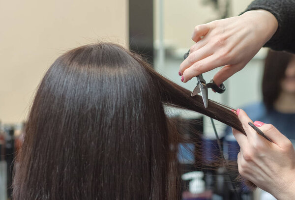 Парикмахер делает стрижку горячими ножницами волос молодой девушке, брюнетке в салоне красоты..