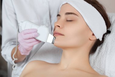 Doktor-kozmetik uzmanı, güzellik salonundaki genç ve güzel bir kadının yüz derisinin ultrason temizleme işlemini yapıyor. Kozmetoloji ve profesyonel cilt bakımı.