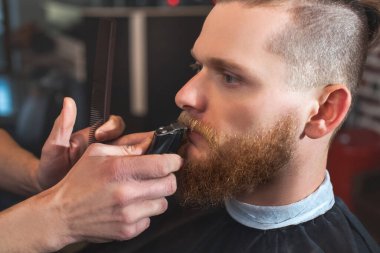 İşteki bir erkek berber, berber dükkanındaki bir adama sakal ve bıyık kesimi yapıyor. Erkek kozmetolojisi, sakal bakımı, şık bir görünüm yaratmak..