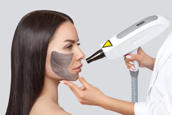A woman with a carbon mask on her face, a beautician does laser carbon peeling on the problem areas of the skin of a young beautiful girl. Cosmetology and professional skin care.