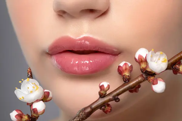 Young girl with beautiful nude make-up and plump lips. Perfect natural lips close up. Near her are beautiful blooming spring sakura flowers. Professional makeup and cosmetology skin care.