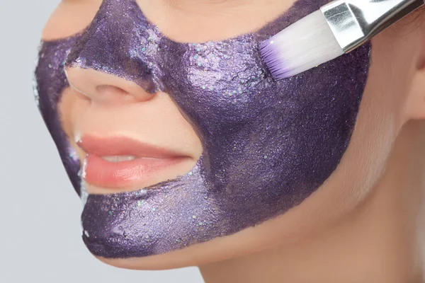 Beautician makes a face mask with purple particles on the face of a woman to rejuvenate the skin. Cosmetology treatment of problem skin on the face and body.